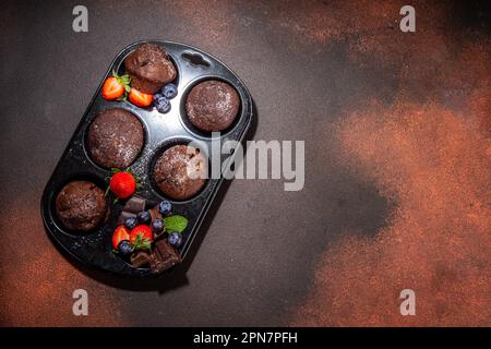 Schokoladen-Muffins mit Beeren. Muffin-Pfanne mit hausgemachten süßen, dunklen Schokoladen-Cupcakes mit Kakaosoße, Zuckerpulver und frischen Erdbeeren, Blaubeeren Stockfoto