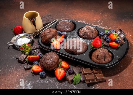 Schokoladen-Muffins mit Beeren. Muffin-Pfanne mit hausgemachten süßen, dunklen Schokoladen-Cupcakes mit Kakaosoße, Zuckerpulver und frischen Erdbeeren, Blaubeeren Stockfoto