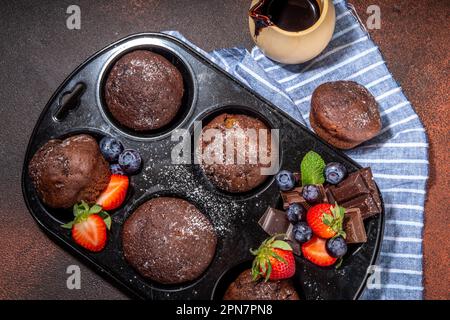 Schokoladen-Muffins mit Beeren. Muffin-Pfanne mit hausgemachten süßen, dunklen Schokoladen-Cupcakes mit Kakaosoße, Zuckerpulver und frischen Erdbeeren, Blaubeeren Stockfoto