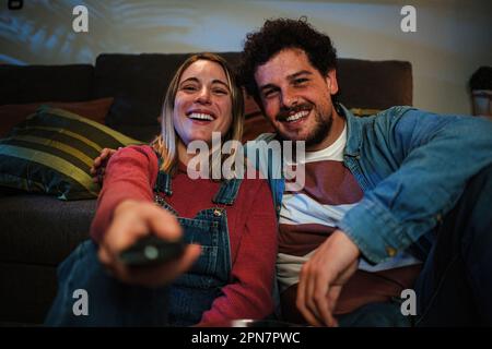 Vorderansicht eines jungen weißlichen Paares, das lächelt und viel Spaß beim Fernsehen hat. Es sitzt zu Hause auf dem Sofa und schaltet den Fernseher mit der Fernbedienung ein. Liebe Beziehung, Mann und Frau. Hochwertiges Foto Stockfoto