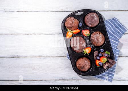 Schokoladen-Muffins mit Beeren. Muffin-Pfanne mit hausgemachten süßen, dunklen Schokoladen-Cupcakes mit Kakaosoße, Zuckerpulver und frischen Erdbeeren, Blaubeeren Stockfoto