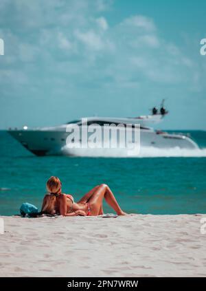 Eine junge Frau im Badeanzug genießt einen sonnigen Tag am Strand und liegt auf einem Boot, während die Wellen sanft gegen das Ufer schlagen Stockfoto