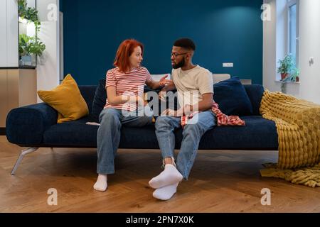 Emotional multiethnisch verärgertes gestresstes Paar, das zu Hause auf der Couch sitzt und sich streitet. Stockfoto