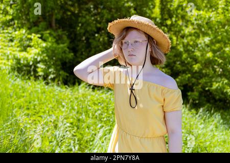 Ein asiatisches Teenager-Mädchen in einem gelben Kleid, eine Brille richtet ihren Hut im Park auf ihren Kopf. Serious Girl mit Hut vor dem Hintergrund von grünem Urlaub Stockfoto