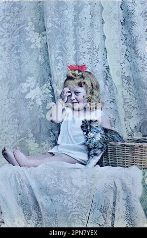 Von einer unzurechenbaren 1904-Postkarte eines kleinen Mädchens mit einer Schleife im Haar, das neben einem Korb mit einer Katze sitzt. Vorhang hinten. Stockfoto