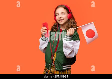 Teenager-Schülerin in lässiger Kleidung und Kopfhörern trägt die kleine japanische Nationalflagge und ein Smartphone. Sprachkurse online. Japanisch Lernen. Liste Stockfoto