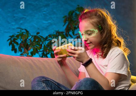 Teenager-Mädchen können zu Hause live streamen, Videospiel-Smartphones in blauem Neonlicht spielen. Junger Gamer, der Online-Spiele-App auf seinem Handy spielt, trägt futuristisches Glas Stockfoto