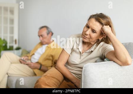 Älterer Mann, der ein Smartphone benutzt, während er sich mit seiner Frau streitet, die sich einsam fühlt und zu Hause auf dem Sofa sitzt Stockfoto