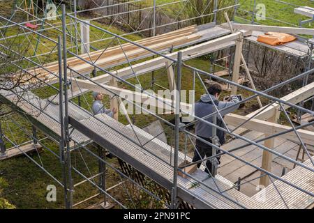 Zwei junge Zimmerleute arbeiten auf einer Baustelle. Ein Zimmermann bohrt mit einem Elektrobohrer ein Loch in einen Sparren und steht auf einem Geländer. Zweiter Zimmermann Stockfoto