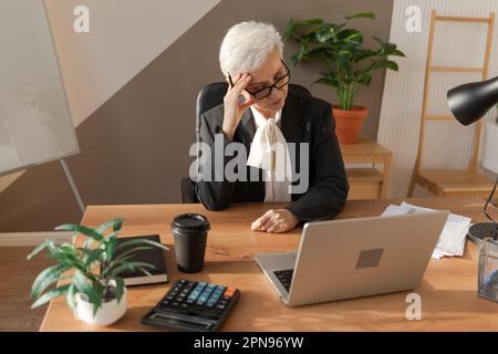 Seniorin berührt Schläfe unter Stress im Büro. Reife Frau müde von der Arbeit Kopfschmerzen krank Schläfen an der Stirn reiben. Computersehsyndrom bei Langnotebooknutzung. Das Office-Syndrom Stockfoto