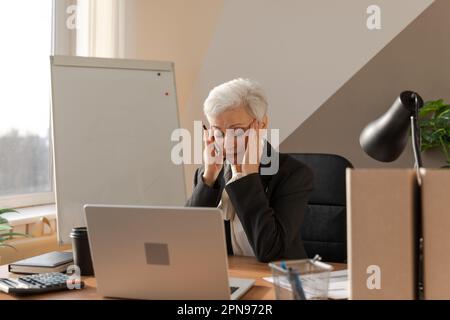 Seniorin berührt Schläfe unter Stress im Büro. Reife Frau müde von der Arbeit Kopfschmerzen krank Schläfen an der Stirn reiben. Computersehsyndrom bei Langnotebooknutzung. Das Office-Syndrom Stockfoto