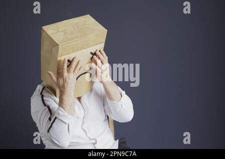 Ein trauriger Mann in einem weißen Hemd mit einer Tasche auf dem Kopf, mit einem gezeichneten traurigen Emoticon, hat Angst und schließt seine Augen mit seinen Händen. Emotionen und Gesten. Stockfoto