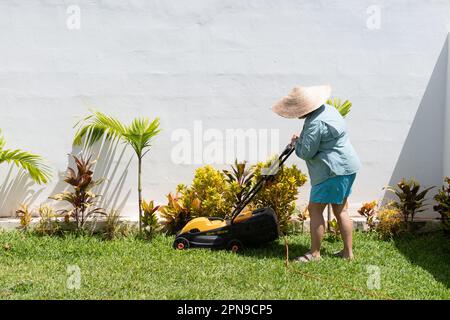 Eine Frau mit blauen Shorts, langärmeligem Hemd und Hut mäht das Gras im Garten ihres Hauses mit einem Rasenmäher. Stockfoto