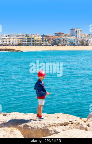 Kleiner Junge mit roter Mütze, der auf einem Felsen vor dem Meer in albufeira, algarve portugal, steht Stockfoto