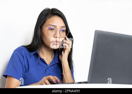 Eine junge Frau arbeitet mit einem Laptop und ruft von einem Mobiltelefon aus an Stockfoto