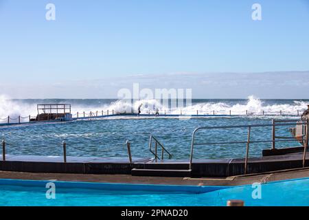 Wellen stürzen über Dee Why Beach Rockpools, Ozeanschwimmbecken. Zwei Jungs in Neoprenanzügen weichen den Wellen aus. Stockfoto