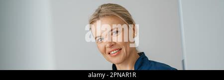 Lächelnder Arzt mit Klemmbrett, der in der Nähe der Rezeption in der Klinikhalle steht und der Kamera schaut Stockfoto