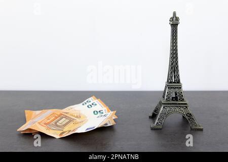 Statue des Eiffelturms aus Metall und Fan von 50-Euro-Scheinen auf schwarzem Tisch vor weißem Wandhintergrund, was einen Hauch von Luxus und Wohlstand vermittelt Stockfoto