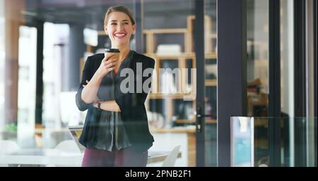 Es geht nichts über eine gute Tasse Kaffee bei der Arbeit. Eine attraktive junge Geschäftsfrau, die allein in ihrem Büro steht und eine Tasse Kaffee hält. Stockfoto