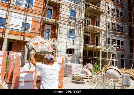 Arbeiter mit Schutzhandschuhen hält den Griff der Schubkarre voll mit zerbrochenem Zellophan, das der Kran auf die Baustelle legt. Im Hintergrund ist unf Stockfoto