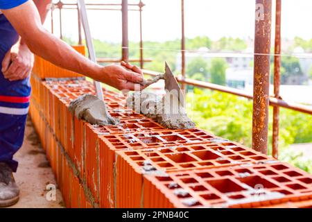 Der Arbeiter verwendet einen Spachtel, eine Kelle, um Mörtel auf rote Blöcke aufzutragen, um Wand und Maurer herzustellen. Das Gebäude befindet sich im Bau. Stockfoto
