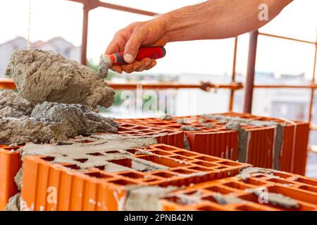 Der Arbeiter verwendet einen Spachtel, eine Kelle, um Mörtel auf rote Blöcke aufzutragen, um Wand und Mauerwerk zu bauen. Das Gebäude ist im Bau und hat ein Gerüst. Stockfoto