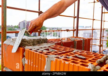 Der Arbeiter verwendet einen Spachtel, eine Kelle, um Mörtel auf rote Blöcke aufzutragen, um Wand und Mauerwerk zu bauen. Das Gebäude ist im Bau und hat ein Gerüst. Stockfoto