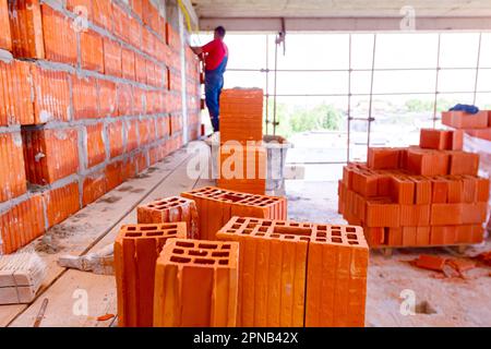 Anhäufung roter Keramikblöcke auf Holzplattform auf der Baustelle. Im Hintergrund baut der Arbeiter Trennwand mit Blöcken und Mörtel. Stockfoto