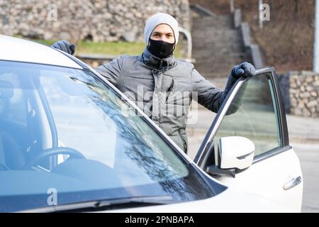 Junge Erwachsene Brünette Mann in chirurgische medizinische Maske, Jacke posiert auf der Straße der Stadt in der Nähe des Autos. Außenaufnahme. Stockfoto