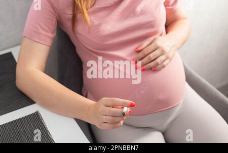 Eine schwangere Frau mit einem großen Bauch sitzt in der Küche am Tisch und raucht. Rauchen und Schwangerschaft. Schaden für die Gesundheit des Kindes und der Mutter, Tobac Stockfoto
