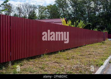 Roter Wellzaun aus Metall rund um den grünen Garten. Die Textur des profilierten Metalls. Stockfoto