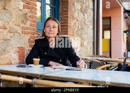 Pensive Frau mittleren Alters in schwarzem Mantel, die mit einer Tasse Kaffee am Tisch sitzt und wegschaut, während sie Notizen auf dem Notizbuch schreibt Stockfoto