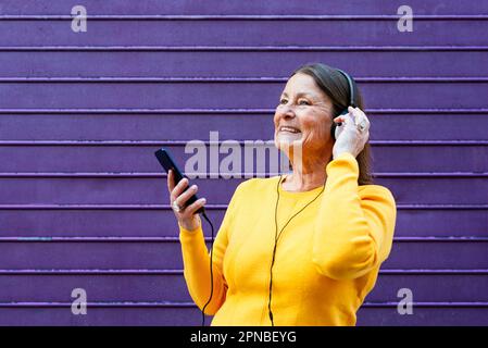 Glückliche Frau in gelbem Pullover, die Musik über Kopfhörer hört und auf dem Handy surft, während sie gegen die violette Wand steht Stockfoto