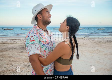 Seitlicher Blick auf lächelnde junge Männer und Frauen, die sich während des Sommerurlaubs in Tulum am Sandstrand in der Nähe des Wellenmeers umarmen Stockfoto