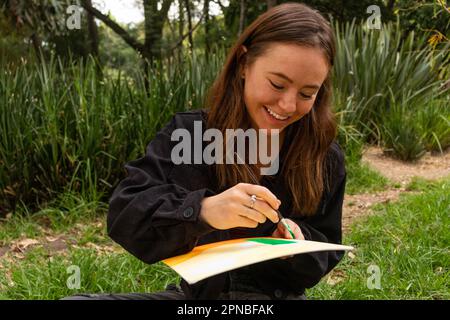 Fröhliche junge Künstlerin, die auf Gras sitzt und mit Pinsel in Papier malt, während sie tagsüber gegen grüne Pflanzen ruht Stockfoto