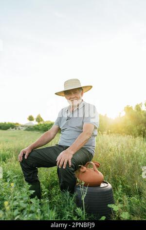 Ein ganzer Körper positiver, bärtiger Bauer in lässiger Kleidung und Strohhut, der auf Baumstamm sitzt und auf der üppigen Wiese auf Su in die Kamera schaut Stockfoto