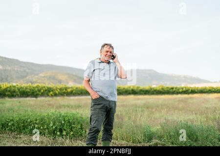 Ein männlicher Bauer mit positivem Bart in lässiger Kleidung, der lächelt und in die Kamera schaut, während er mit dem Mobiltelefon in der Nähe der grünen Wiese in Mount steht Stockfoto
