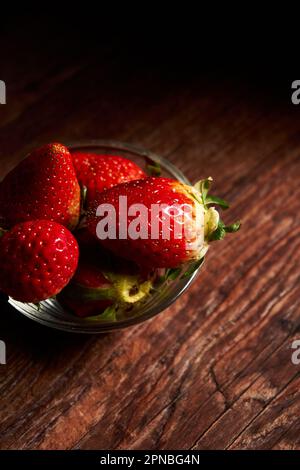 Köstliche frische Erdbeeren mit grünen Blättern in einer transparenten Schüssel auf einem Holztisch Stockfoto