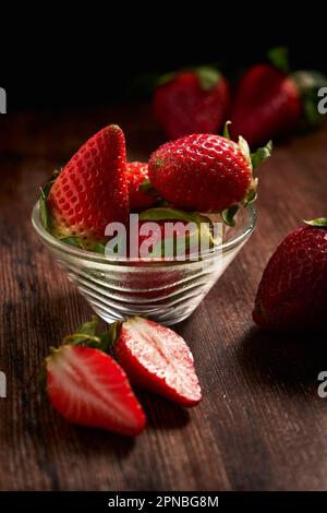 Köstliche frische Erdbeeren mit grünen Blättern in einer transparenten Schüssel auf einem Holztisch Stockfoto