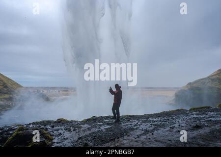 Seitenansicht des männlichen Reisenden, der auf einer felsigen Klippe steht und mit dem Smartphone Fotos macht, während er Zeit in der Nähe des fließenden Seljalandsfoss-Wasserfalls verbringt Stockfoto