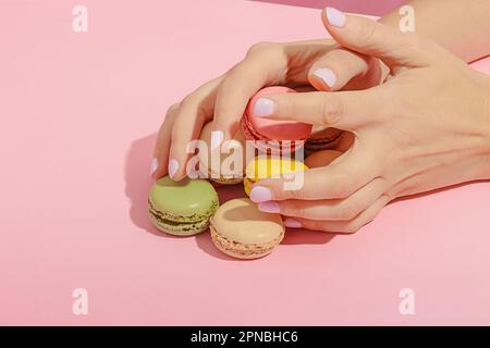 Unbekannte Frau mit Maniküre, die einen Haufen leckerer, süßer, mehrfarbiger Makronen von der pinkfarbenen Oberfläche nimmt Stockfoto