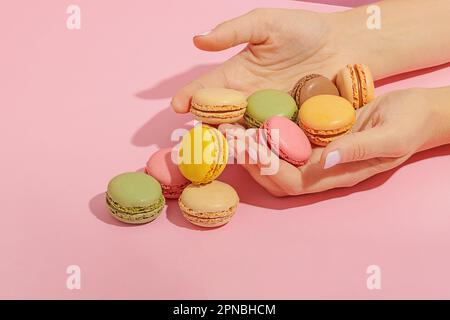 Unbekannte Frau mit Maniküre, die einen Haufen leckerer, süßer, mehrfarbiger Makronen von der pinkfarbenen Oberfläche nimmt Stockfoto