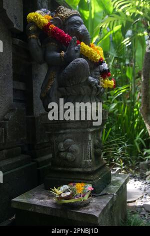 Traditionelle Ganesha-Statue mit Blumenkranz im balinesischen Garten. Lord Ganesha soll Glück bringen, und so wird er vor allem verehrt Stockfoto