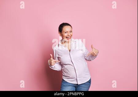 Eine reizende schwangere Frau, die Aufregung ausdrückt, das erste Baby tritt, mit dem Finger auf ihren Bauch zeigt, vor der Kamera lächelt Stockfoto