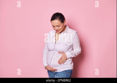 Eine reizende, glückliche schwangere Frau, die süß lächelt, während sie ihren Bauch berührt und streichelt, und ihre sorgenfreie Schwangerschaft genießt Stockfoto