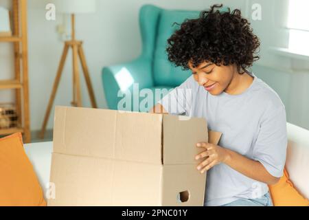 Afrikanisches Mädchen, das die Lieferung auspackt und in die Schachtel schaut. Glückliche Frau, die eine Schachtel öffnet. Eine Frau bekommt ein Paket, das sich die gelieferten Waren ansieht. Zufriedene Kunden positives Feedback, schnelle Expresslieferung Stockfoto