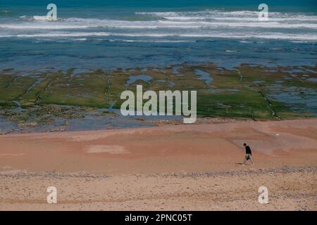 Sidi Ifni, Marokko - aus der Vogelperspektive kann man am Strand spazieren gehen. Brauner orangefarbener Sand, felsige Küste mit grünem Moos, blaue Wellen des Atlantischen Ozeans. Stockfoto