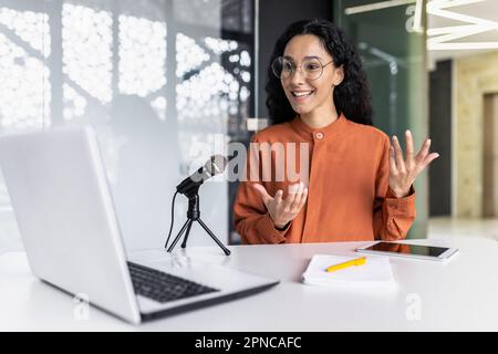 Erfolgreiche lateinamerikanische Bloggerin, die in modernen Büros Online-Video-Podcasts aufnimmt, weibliche Mitarbeiterin, die ein professionelles Mikrofon für Online-Stream und Laptop verwendet, lächelt und Gesten macht. Stockfoto