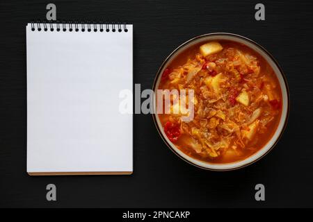 Hausgemachter Kohl und Kartoffelsuppe in einer Schüssel, leeres Notizbuch auf schwarzem Hintergrund, Draufsicht. Über Kopf, von oben, flach verlegt. Stockfoto