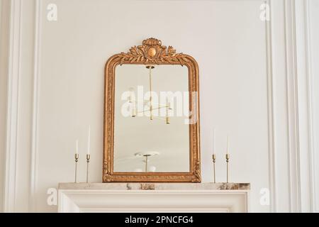 Ein Spiegel in einem klassischen Rahmen auf dem Kaminsims neben Kerzenleuchtern mit Kerzen. Teil des Innenraums mit einer Reflexion des Kronleuchters an der Decke. Klassisches Interieur mit Kopierraum. Stockfoto